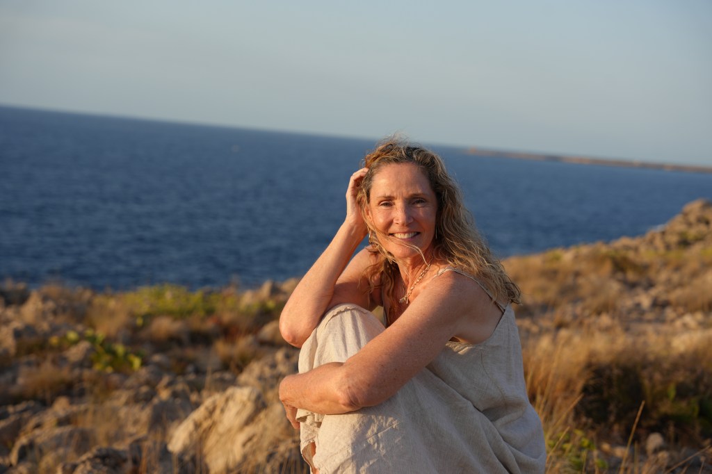 A smiling woman with long, wavy hair sitting on rocks by the ocean, wearing a light-colored outfit, with a scenic seascape in the background.